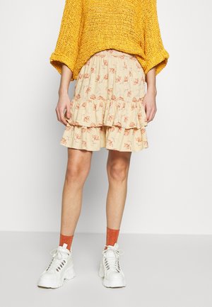 Jupe plissée en beige à motifs floraux avec volants, fabriquée en tissu léger. Associée à des baskets blanches à semelles épaisses et des chaussettes orange.