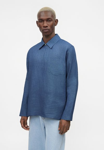 Άνδρας με κοντά ξανθά μαλλιά φοράει ένα μακρυμάνικο μπλε υφασμάτινο πουκάμισο και ανοιχτό μπλε τζιν, στέκεται μπροστά σε απλό λευκό φόντο.