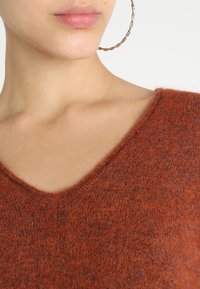 Pull en laine marron avec un col en V, présentant un tissu doux et texturé. Gros plan sur le col et la zone de l'épaule.