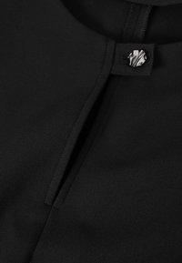Gros plan sur un tissu noir avec un col en forme de trou de serrure, fermé par un petit bouton rond argenté texturé.