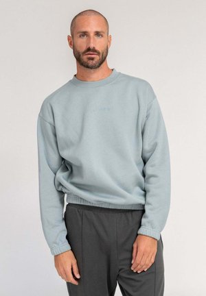 Man with short hair and beard wearing light blue sweatshirt and dark gray pants, standing against plain light background.