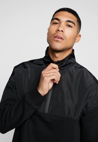 Pullover nero con colletto a zip a quarto, una combinazione di materiali opachi e lucidi, e polsini a coste. Design semplice con accenti minimi.