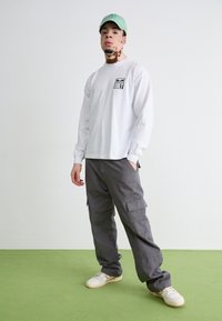 Long-sleeve white shirt with a black "OBEY" logo, gray cargo pants with side pockets, green cap, and cream sneakers on a green floor.