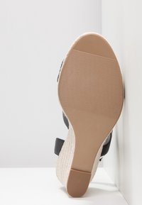 Semelle d'une sandale compensée beige avec semelle en caoutchouc texturée et bride noire à la cheville sur un fond d'angle blanc.