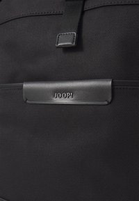 Mochila de tela negra texturizada con un acento de cuero que lleva grabado "JOOP!", mostrando un diseño elegante y materiales duraderos.