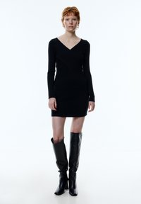 Schwarzes, langärmliges Mini-Kleid mit V-Ausschnitt und diagonalen Nahtdetails, kombiniert mit schwarzen, stiefellangen Lederstiefeln. Glatte Textur.