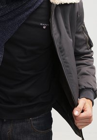 Harmaa pommikennys takki, jossa on karvakaulus, vetoketjulliset taskut ja ribbireunat, yllä musta pitkähihaisten paita ja tummat farkut.