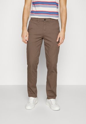 Man draagt bruine rechte broek, witte sneakers en een wit T-shirt met rode en blauwe horizontale strepen.