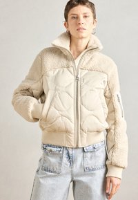 Beige steppelt kabát műszőrme díszítéssel, magas gallérral, bordázott mandzsettákkal és cipzáras elülső résszel. Világos farmernadrággal párosítva.