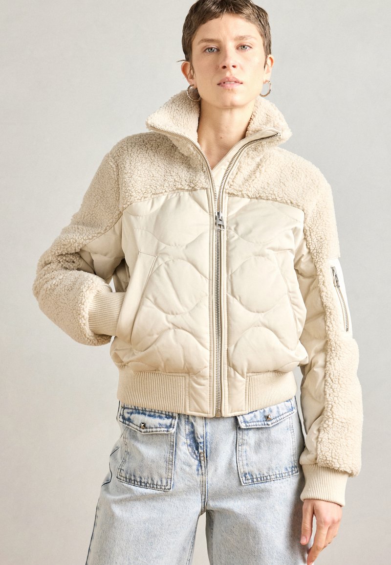 Beige steppelt kabát műszőrme díszítéssel, magas gallérral, bordázott mandzsettákkal és cipzáras elülső résszel. Világos farmernadrággal párosítva.