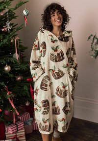 Oversize cremefarvet fleece-hoodie med leopard- og kristtornprint; har hætte og lommer, båret over et let outfit, ved siden af et juletræ.