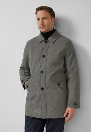 Mann trägt grauen, geknöpften Mantel über schwarzem Rollkragenpullover, Hände in den Taschen, steht vor einer hellgrauen Wand und schaut leicht nach rechts.