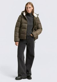 Vadderad hujacka i olivgrön färg med vit fleecekrage och quiltad design. Kombinerad med mörka jeans med vida ben och svarta stövlar.