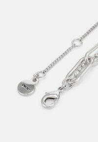 Sølvmetal kæde med en lås og et cirkulært DKNY-logo skilt. Har en glat tekstur, metallisk glans og sammenkoblede designelementer.