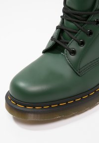 Zielony skórzany botek za kostkę z czarnymi sznurowadłami, okrągłym noskiem i żółtym przeszyciem wzdłuż podeszwy. Gruba, przezroczysta gumowa podeszwa z detaliami bieżnika.