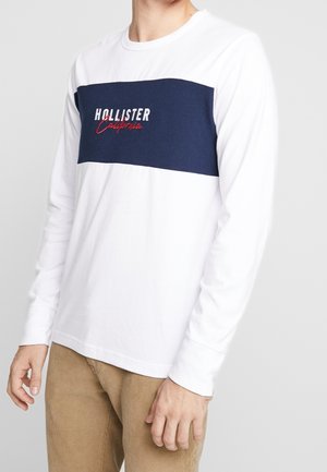 Mężczyzna w białej koszuli z długim rękawem z granatowym panelem i napisem "Hollister California", w połączeniu z jasnobrązowymi spodniami.