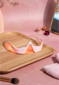 Witte en heldere gemusterde zonnebril met een geometrisch ontwerp, rustend op een houten schaal tegen een roze hexagonaal achtergrond.