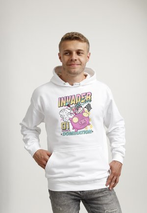 Junger Mann trägt weißen Hoodie mit buntem Cartoon-Schwein, karierten Fahnen und dem Text „Invader 01 World Domination“ auf der Vorderseite, Hände in den Taschen.