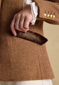 Main avec une bague en argent tirant un mouchoir de poche marron à motifs de la manche d'un blazer marron en chevrons avec des boutons dorés et un poignet blanc.