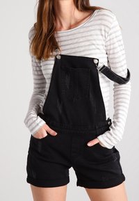 Black denim overalls with distressed details, featuring adjustable straps and a front pocket, paired with a gray striped long-sleeve shirt.