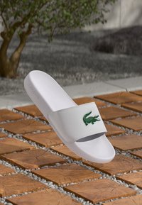 Hvid slide sandal med en tekstureret strop, prydet med et grønt krokodille-logo. Glat plastmateriale, flad sål, minimalistisk design.