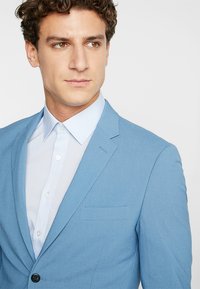 Chaqueta de traje azul claro en una tela suave, con cierre de un solo botón, solapas de muesca y un bolsillo en el pecho. Camisa blanca de botones debajo.