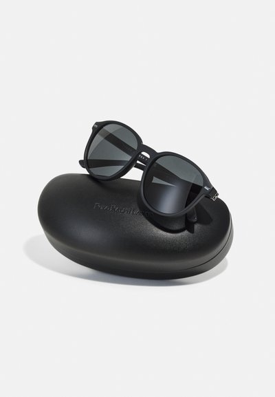 Melnas saulesbrilles ar pelēkiem stikliem, ar apaļu formu un matētu apdari, atpūšas uz melnas ovālas cietās kārbas.