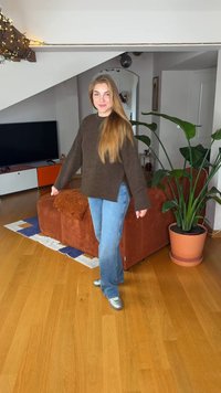 Brun stickad tröja, ljusblå jeans och silvriga skor. Står i ett modernt rum med soffa, växt och trägolv.