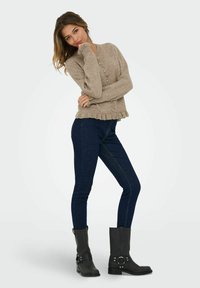 Bežs kardigāns ar pogu aizdari un rējamas malām, apvienots ar tumši zilām šaurām džinsām un melniem potītes zābakiem ar sudraba akcentiem.