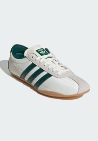 Bijele tenisice s zelenim prugama, sueded prednji deo, gumeni đon i zeleni logo na jeziku. Distinktivan retro dizajn.