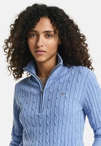 Helles blauer Kabelstrickpullover mit einem Reißverschlusskragen und subtilen Logodetails. Weicher Stoff und taillierte Passform, geeignet zum Schichten.