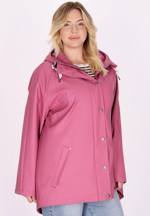 Kobieta o blond włosach ubrana w różowy pelerynkę przeciwdeszczową z kapturem na paskowanym T-shircie i jasnoniebieskich dżinsach, stojąca na jednolitym tle.