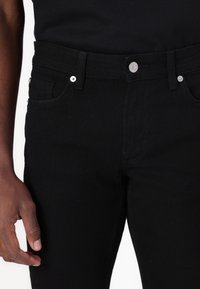 Detalles en primer plano de una persona que lleva jeans negros y una camisa negra, mostrando la cintura, los bolsillos, el botón y una mano relajada al lado.