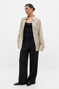 Šviesiai rudas langeliais marškinis su ilgomis rankovėmis, laisvu siluetu ir sagčių uždarymu, dėvimas ant juodos maikės ir derinamas su plačiomis juodomis kelnėmis.