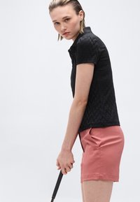 Νεαρή γυναίκα με μπαστούνι του γκολφ, φοράει μαύρο πόλο με υφή και ροζ σορτς σαλιμοντί, μπροστά σε απλό λευκό φόντο.