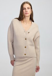 Cardigan beige a maglia con scollatura a V, orlo a coste e bottoni dorati. Presenta una vestibilità rilassata e una texture morbida, abbinato a una gonna coordinata.