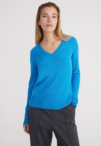 Pullover V-neck blu brillante realizzato in materiale morbido con polsini e orlo a coste. Indossato con pantaloni grigi scuri, la texture appare liscia.