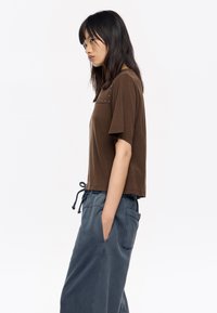Camiseta marrón cortada con mangas cortas, con acentos de tachuelas en los hombros. Llevada con pantalones azul con cordón, bolsillo lateral visible.