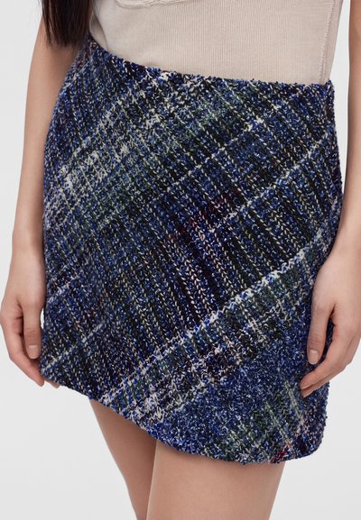 Gros plan d'une mini-jupe écossaise texturée bleue et verte avec un motif diagonal, portée avec un haut beige.