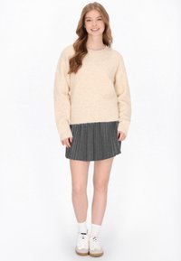 Strickpullover in cremigem Beige mit weicher Textur, kombiniert mit einem dunkelgrauen Faltenrock, getragen mit weißen Socken und beigen Turnschuhen.