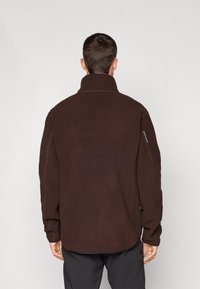 Smeđi flis pulover s visokim ovratnikom, dugim rukavima i rebrastim manžetama. Ima zipped džep na lijevom rukavu. Meka, teksturirana površina.