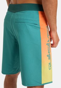 Bañador en color turquesa con un panel de degradado en naranja y amarillo. Cuenta con un bolsillo en la parte trasera y el logo "Quiksilver" en un color en contraste en el lateral.