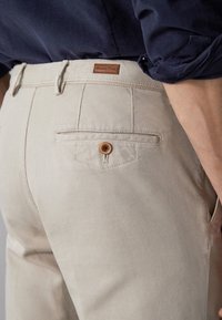 Beige bomullbyxor med en strukturerad yta, med en bakficka med en brun knapp och en liten läderetikett ovanför fickan.