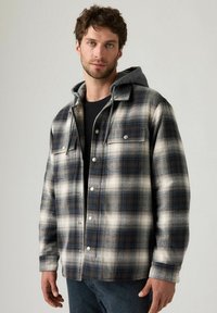 Camicia flanella a quadri in tonalità di nero, bianco e blu, con cappuccio grigio staccabile e chiusura con bottoni a pressione. Tessuto testurizzato con tasche.