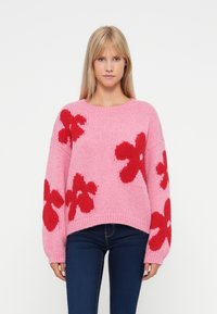 Mulher loira vestindo um suéter de malha rosa com grandes padrões de flores vermelhas e jeans escuros, de pé contra um fundo branco.
