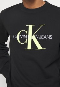 Černá mikina se žebrovaným výstřihem, se velkým neonově žlutým grafickým prvkem "CK" a bílým textem "CALVIN KLEIN JEANS". Měkká tkanina.