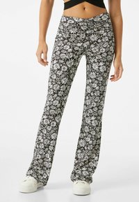 Pantalones negros con estampado de margaritas blancas, ajustados en la cintura y acampanados en la parte inferior. Hechos de tela ligera y combinados con zapatillas blancas.