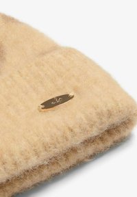 Khaki värvi kootud müts pehmest villast, tekstureeritud viimistlusega, külje peal metallist logo plaadiga.