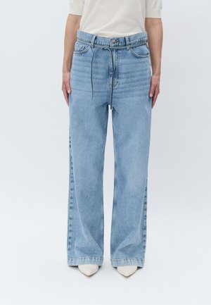 Persona che indossa jeans a vita alta color azzurro chiaro con gamba larga, cintura con coulisse e scarpe bianche appuntite, in piedi davanti a uno sfondo semplice.