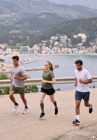 Drei Läufer auf einem Küstenweg: einer in einem grauen Shirt und Shorts, einer in einem grünen Oberteil und schwarzem Rock und einer in einem weißen Shirt und blauen Shorts.
